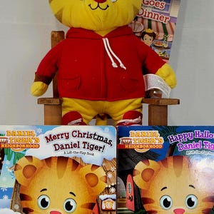 Peut inclure: Un jouet en peluche Daniel Tiger jaune portant un sweat &agrave; capuche rouge et un pantalon jaune, assis sur une petite chaise en bois. Trois livres pour enfants sont devant le jouet, dont "Joyeux No&euml;l, Daniel le Tigre !"
