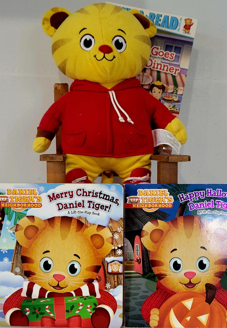 Peut inclure: Un jouet en peluche Daniel Tiger jaune portant un sweat &agrave; capuche rouge et un pantalon jaune, assis sur une petite chaise en bois. Trois livres pour enfants sont devant le jouet, dont "Joyeux No&euml;l, Daniel le Tigre !"