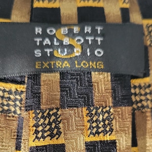 May include: A close-up of a Robert Talbott Studio extra long tie. The tie features a repeating pattern of black, gold, and brown squares. The label is black with gold lettering.