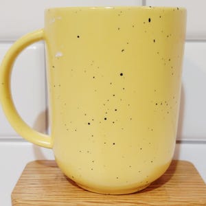 May include: A yellow ceramic mug with black speckles and a curved handle. The mug is sitting on a small, light-colored wooden coaster. The background is a white tiled wall.