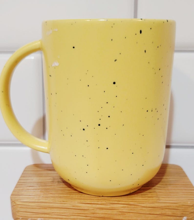 May include: A yellow ceramic mug with black speckles and a curved handle. The mug is sitting on a small, light-colored wooden coaster. The background is a white tiled wall.