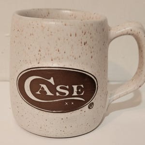 May include: A ceramic mug with a speckled off-white finish and a brown handle. The front features a brown oval logo containing the white text CASE with two small stars underneath and a registered trademark symbol to the right of the oval design.