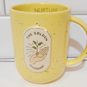 May include: Yellow ceramic mug with black speckles and a curved handle. The mug features an oval design with the words "THE GOLDEN GARDENER" around a hand holding a sprouting plant. The word "NURTURE" is printed on the rim.