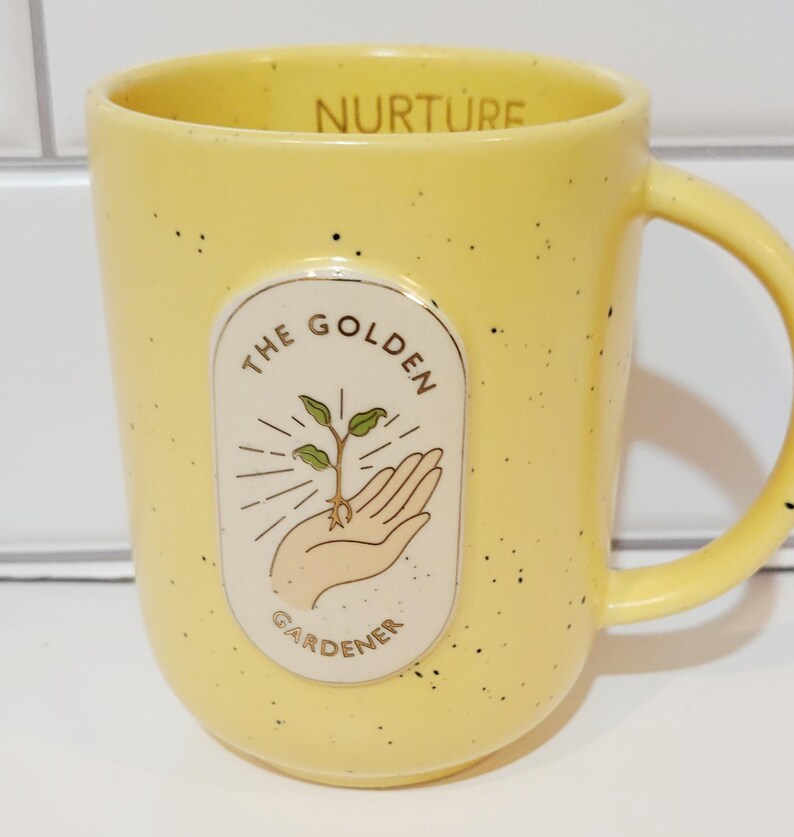 May include: Yellow ceramic mug with black speckles and a curved handle. The mug features an oval design with the words "THE GOLDEN GARDENER" around a hand holding a sprouting plant. The word "NURTURE" is printed on the rim.