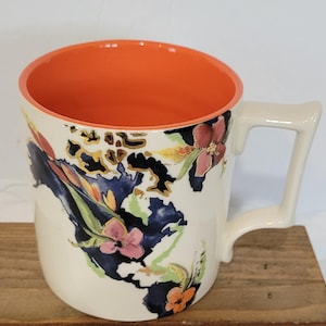 Pu&ograve; includere: Tazza in ceramica color crema con interno arancione e un manico unico. La tazza &egrave; decorata con un motivo floreale in rosa, arancione e blu, con dettagli dorati. La tazza &egrave; appoggiata su una superficie di legno.