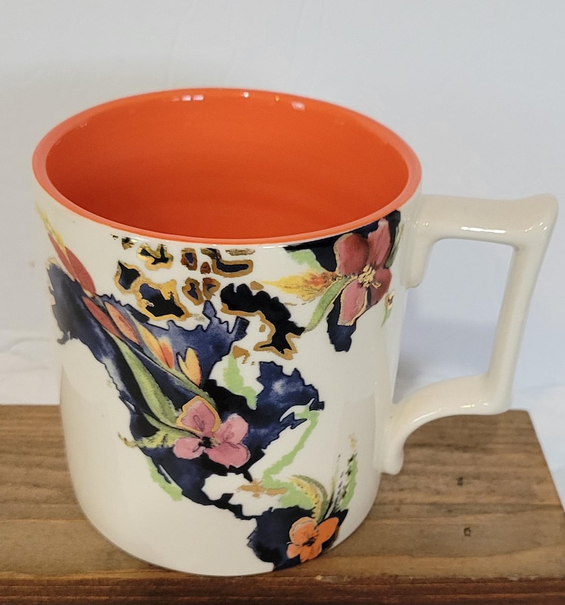 Pu&ograve; includere: Tazza in ceramica color crema con interno arancione e un manico unico. La tazza &egrave; decorata con un motivo floreale in rosa, arancione e blu, con dettagli dorati. La tazza &egrave; appoggiata su una superficie di legno.