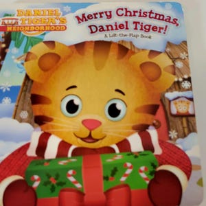 Peut inclure: Couverture de livre pour enfants "Joyeux No&euml;l, Daniel le Tigre !" avec un tigre de dessin anim&eacute; tenant un cadeau de No&euml;l. Le livre fait partie de la s&eacute;rie "Daniel le Tigre" et est un livre &agrave; rabats. Le tigre porte une &eacute;charpe rouge et blanche.