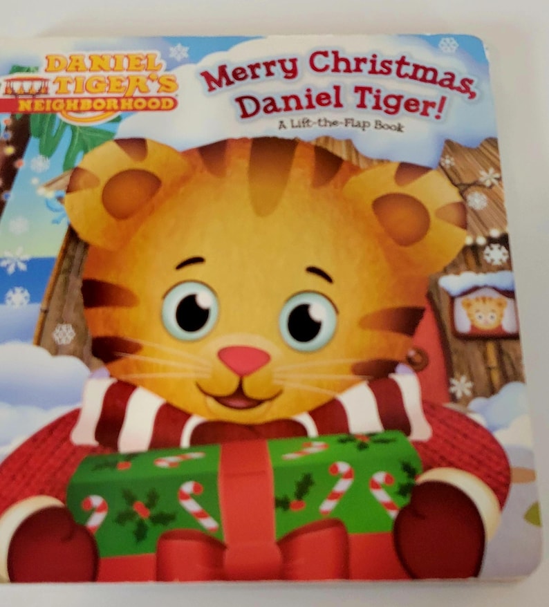 Peut inclure: Couverture de livre pour enfants "Joyeux No&euml;l, Daniel le Tigre !" avec un tigre de dessin anim&eacute; tenant un cadeau de No&euml;l. Le livre fait partie de la s&eacute;rie "Daniel le Tigre" et est un livre &agrave; rabats. Le tigre porte une &eacute;charpe rouge et blanche.
