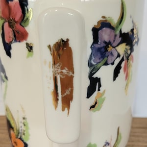 Pu&ograve; includere: Una tazza di ceramica bianca con un vivace motivo floreale con fiori rosa, viola e arancioni. Il bordo interno &egrave; arancione e il manico ha un design strutturato marrone e oro. La tazza &egrave; appoggiata su una superficie di legno.