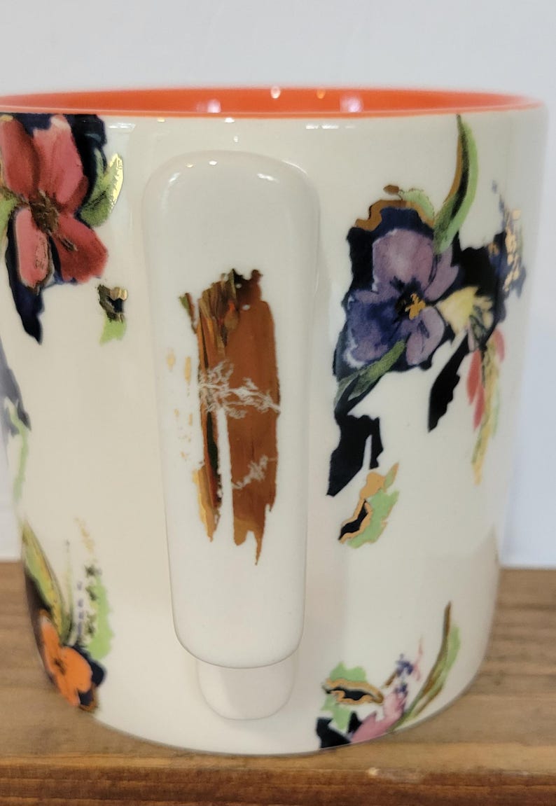 Pu&ograve; includere: Una tazza di ceramica bianca con un vivace motivo floreale con fiori rosa, viola e arancioni. Il bordo interno &egrave; arancione e il manico ha un design strutturato marrone e oro. La tazza &egrave; appoggiata su una superficie di legno.