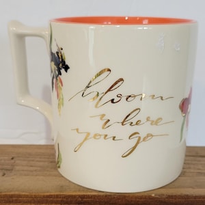 Pu&ograve; includere: Tazza in ceramica color crema con interno arancione e manico curvo. La tazza presenta una scritta dorata che recita "Bloom where you go" e illustrazioni floreali in rosso, nero e verde.