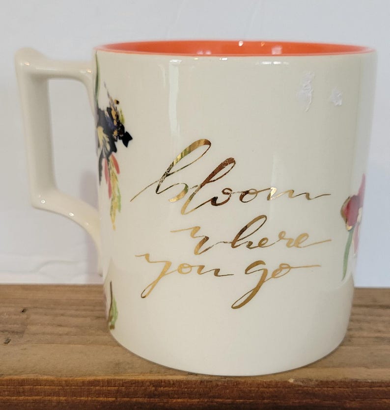 Pu&ograve; includere: Tazza in ceramica color crema con interno arancione e manico curvo. La tazza presenta una scritta dorata che recita "Bloom where you go" e illustrazioni floreali in rosso, nero e verde.