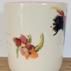 Pu&ograve; includere: Una tazza in ceramica color crema con disegni floreali. I fiori sono nelle tonalit&agrave; del rosso, dell'arancione e del blu, con accenti dorati. Il bordo della tazza &egrave; di colore rosso-arancio.