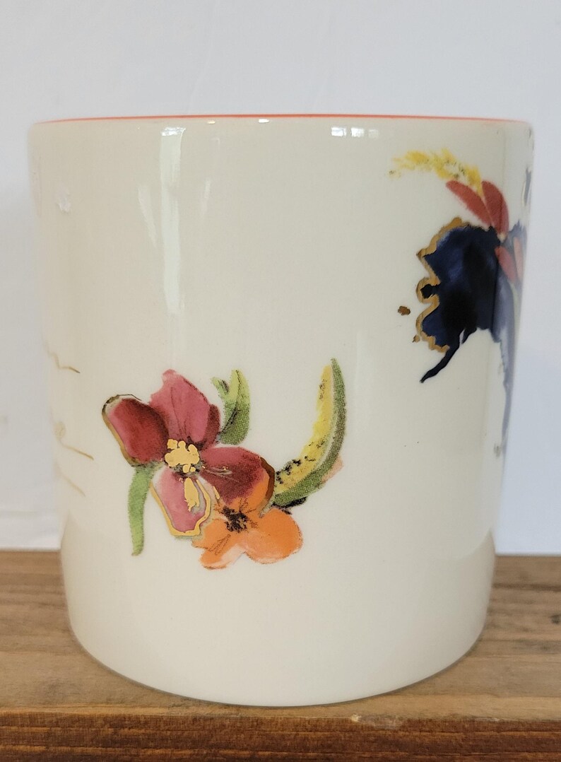 Pu&ograve; includere: Una tazza in ceramica color crema con disegni floreali. I fiori sono nelle tonalit&agrave; del rosso, dell'arancione e del blu, con accenti dorati. Il bordo della tazza &egrave; di colore rosso-arancio.