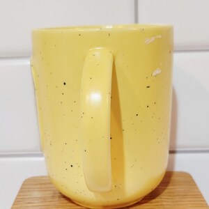 May include: A yellow ceramic mug with a rounded base and a large handle. The mug has small black speckles and sits on a wooden coaster. The background is a white tiled wall.