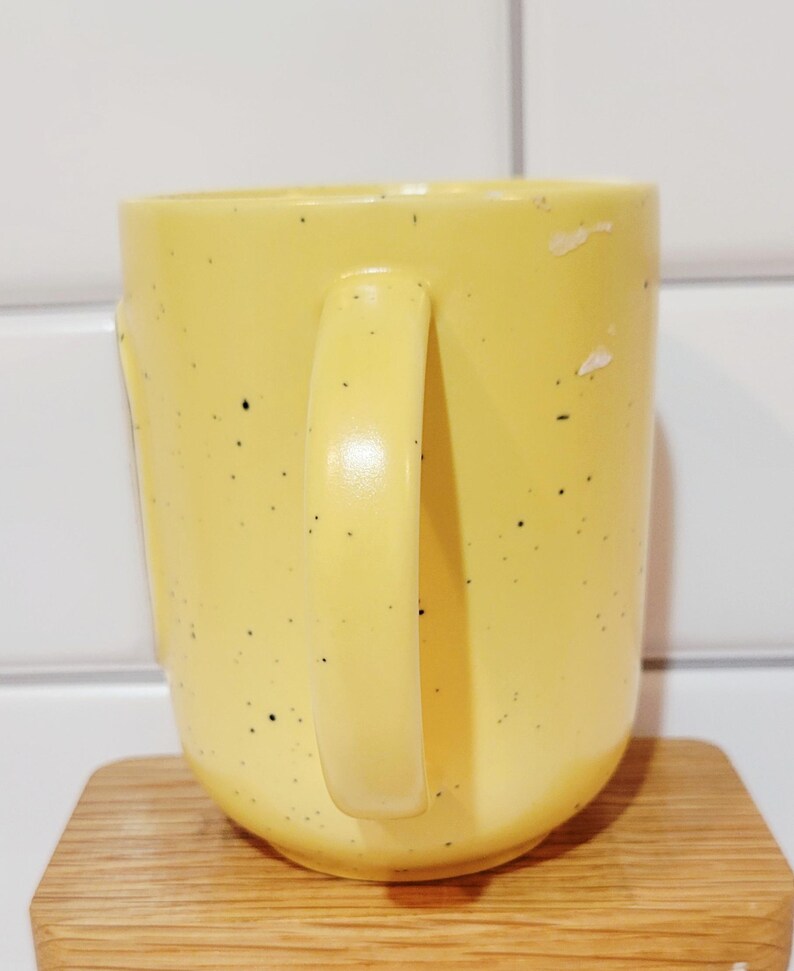 May include: A yellow ceramic mug with a rounded base and a large handle. The mug has small black speckles and sits on a wooden coaster. The background is a white tiled wall.