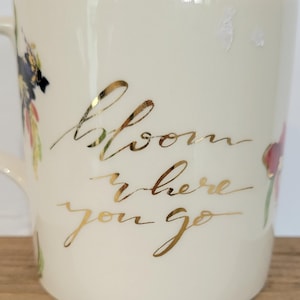 Pu&ograve; includere: Tazza in ceramica color crema con scritta dorata "Bloom where you go". La tazza presenta disegni floreali nei toni del rosa, viola e verde. Il bordo della tazza ha un accento color pesca.