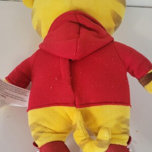 Peut inclure: Une peluche repr&eacute;sentant un tigre, vue de dos, portant une veste &agrave; capuche rouge et un pantalon jaune &agrave; rayures marron. Le tigre a des oreilles jaunes et une queue ray&eacute;e. Des chaussures rouges et blanches compl&egrave;tent la tenue.