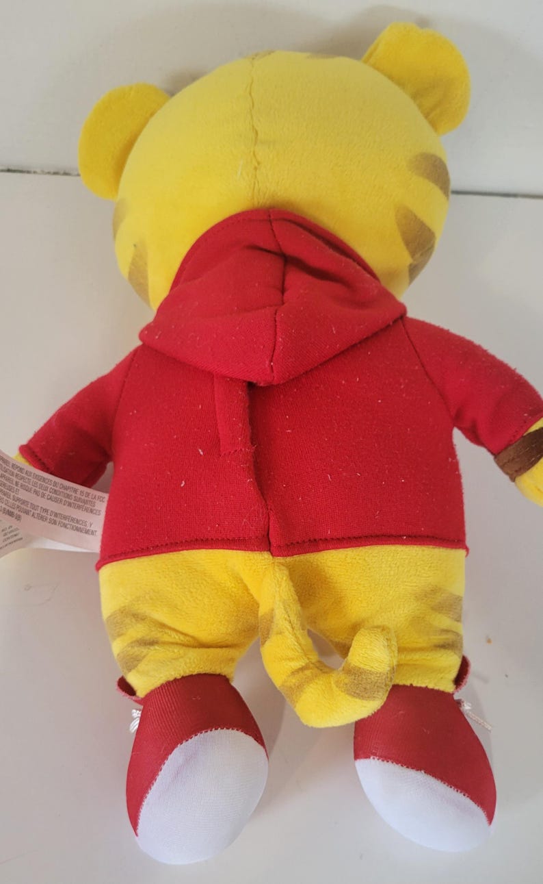 Peut inclure: Une peluche repr&eacute;sentant un tigre, vue de dos, portant une veste &agrave; capuche rouge et un pantalon jaune &agrave; rayures marron. Le tigre a des oreilles jaunes et une queue ray&eacute;e. Des chaussures rouges et blanches compl&egrave;tent la tenue.