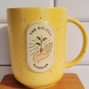May include: Yellow ceramic mug with a speckled finish. The mug features a cream-colored oval emblem with the words "THE GOLDEN GARDENER" and an illustration of a hand holding a sprouting plant. The handle is a matching yellow.