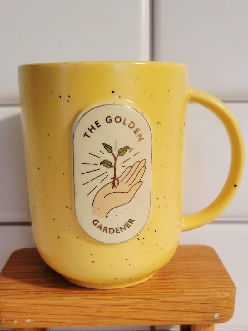 May include: Yellow ceramic mug with a speckled finish. The mug features a cream-colored oval emblem with the words "THE GOLDEN GARDENER" and an illustration of a hand holding a sprouting plant. The handle is a matching yellow.