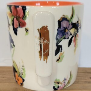 Pu&ograve; includere: Tazza in ceramica bianca con un design floreale con fiori rossi, viola e arancioni. L'interno della tazza &egrave; arancione e il manico ha un accento marrone e dorato. La tazza &egrave; su una superficie di legno.