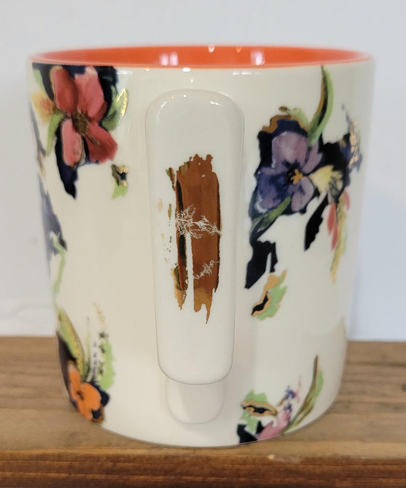 Pu&ograve; includere: Tazza in ceramica bianca con un design floreale con fiori rossi, viola e arancioni. L'interno della tazza &egrave; arancione e il manico ha un accento marrone e dorato. La tazza &egrave; su una superficie di legno.