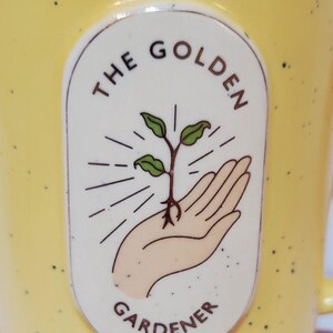 May include: Yellow ceramic mug with black speckles. The mug features an oval design with the words "THE GOLDEN GARDENER" surrounding an illustration of a hand holding a sprouting plant. The mug has a handle.