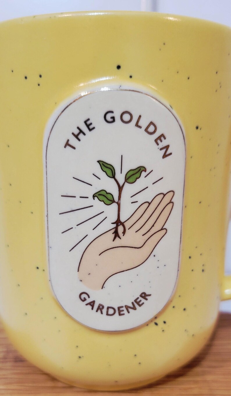 May include: Yellow ceramic mug with black speckles. The mug features an oval design with the words "THE GOLDEN GARDENER" surrounding an illustration of a hand holding a sprouting plant. The mug has a handle.