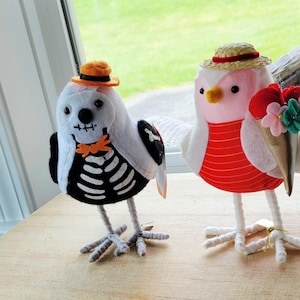 May include: Two decorative bird figurines. One is white with a skeleton design, a black wing, and an orange hat. The other is pink and white with a red striped shirt, a straw hat, and a bouquet of flowers.