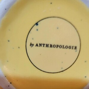 May include: Close-up of a yellow ceramic surface with a speckled pattern and the text "by ANTHROPOLOGIE" inside a black-lined circle. The background is a soft yellow.