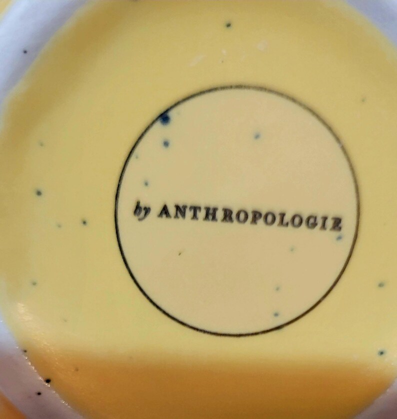 May include: Close-up of a yellow ceramic surface with a speckled pattern and the text "by ANTHROPOLOGIE" inside a black-lined circle. The background is a soft yellow.