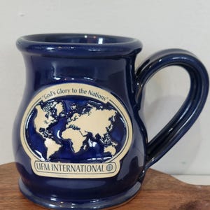 May include: A navy blue ceramic mug with a large handle. The mug features a world map design in white, with the text "God's Glory to the Nations" and "UFM INTERNATIONAL".