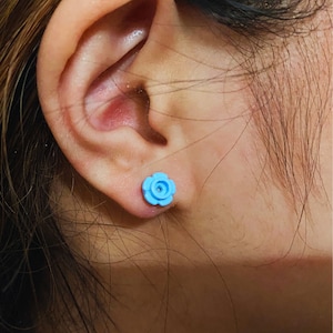 Pode incluir: Um close-up de uma orelha de uma pessoa com um brinco de botão em forma de flor azul claro.