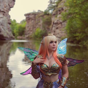 May include: A woman dressed as a fairy with iridescent wings stands in a forest by a river. She is wearing a gold and green bodice, a light blue skirt, and a crown.