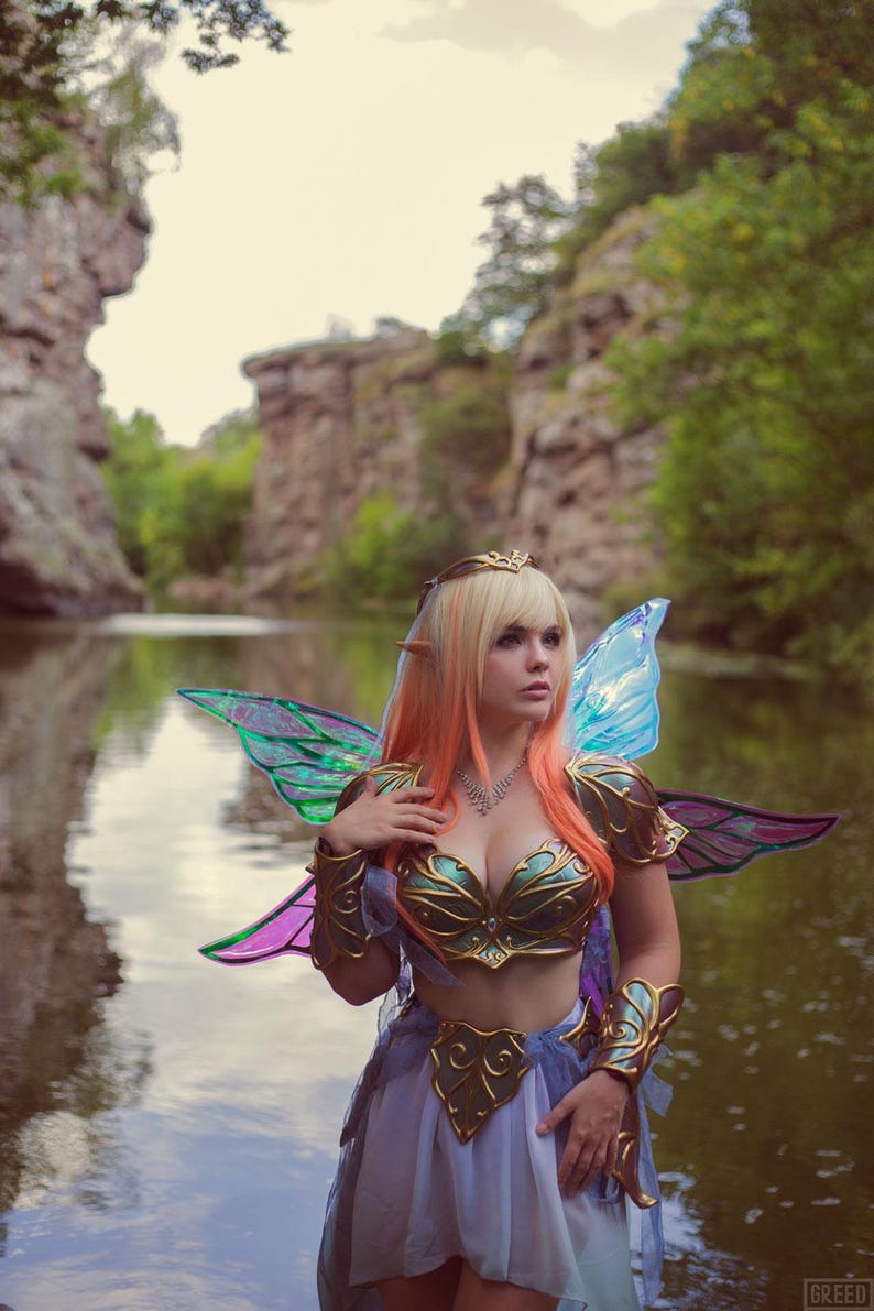 May include: A woman dressed as a fairy with iridescent wings stands in a forest by a river. She is wearing a gold and green bodice, a light blue skirt, and a crown.