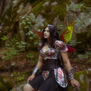 May include: A woman in a fantasy costume with iridescent wings, a silver and red metal breastplate, and a black skirt. She is wearing a silver crown and has long dark hair. She is standing in a forest setting.
