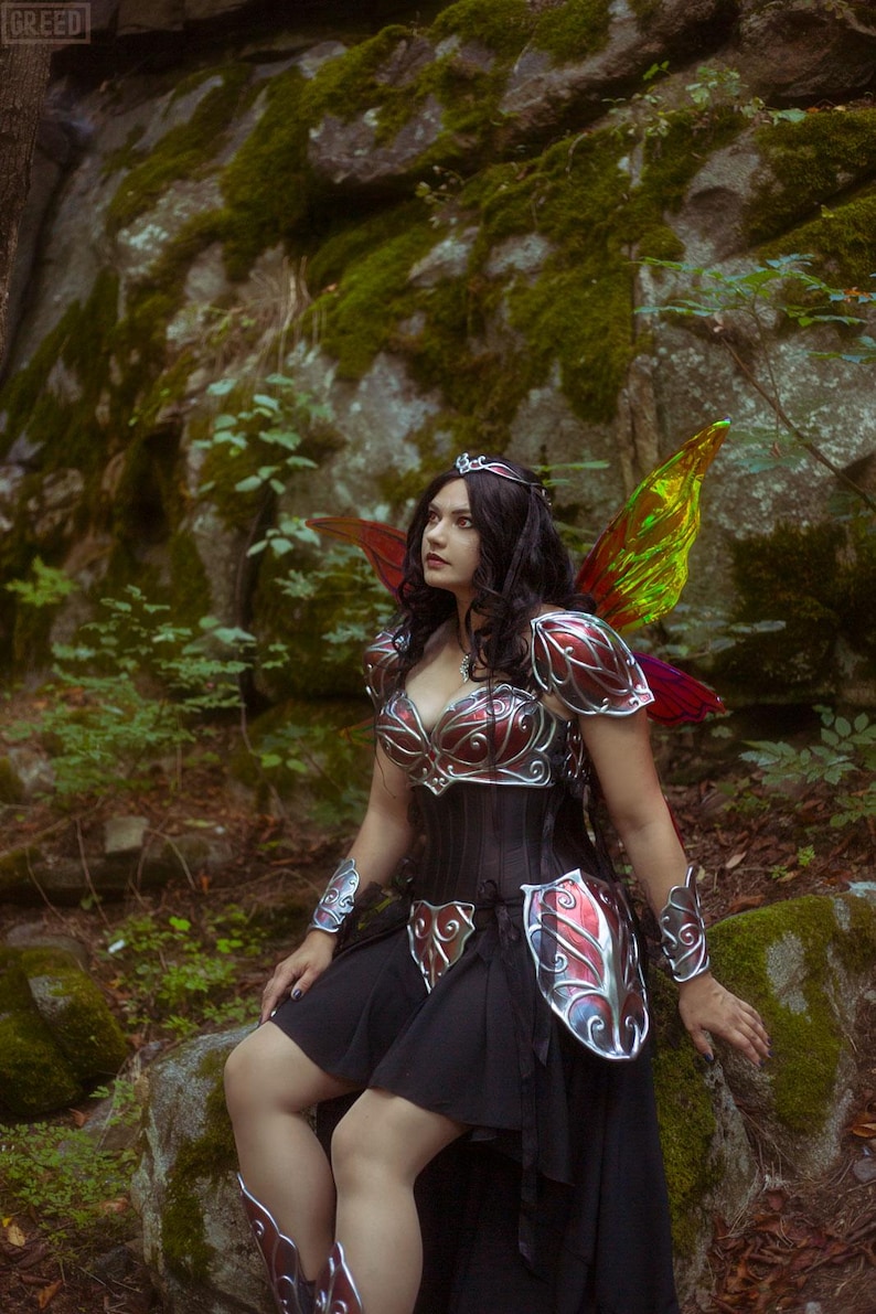 May include: A woman in a fantasy costume with iridescent wings, a silver and red metal breastplate, and a black skirt. She is wearing a silver crown and has long dark hair. She is standing in a forest setting.