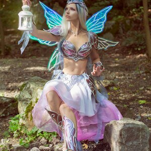 May include: A woman dressed as a fairy with iridescent blue wings, a silver and pink bodice, and a flowing pink skirt. She is holding a lantern and sitting on a rock in a forest setting.