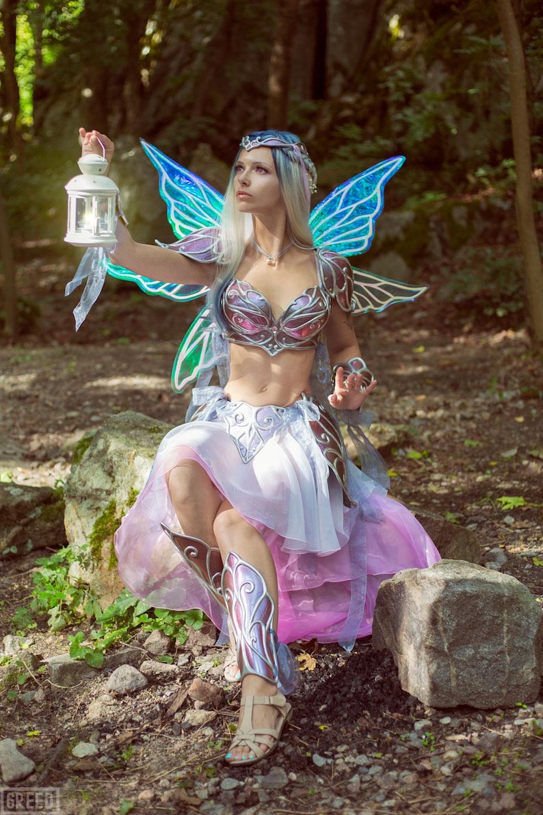 May include: A woman dressed as a fairy with iridescent blue wings, a silver and pink bodice, and a flowing pink skirt. She is holding a lantern and sitting on a rock in a forest setting.