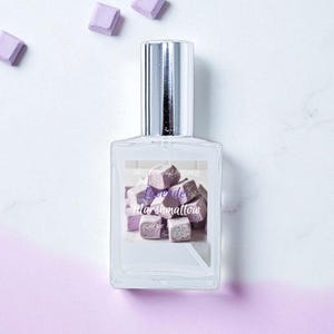 May include: A clear glass perfume bottle with a silver cap, featuring a label with the text "Lune de Marshmallow" over a graphic of purple marshmallow cubes. Several marshmallow cubes are scattered around the bottle on a white and purple background.