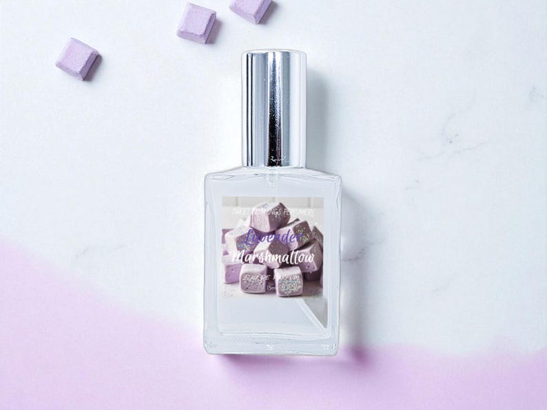 May include: A clear glass perfume bottle with a silver cap, featuring a label with the text "Lune de Marshmallow" over a graphic of purple marshmallow cubes. Several marshmallow cubes are scattered around the bottle on a white and purple background.