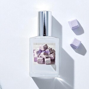 May include: A clear perfume bottle with a silver cap and a label featuring a graphic of purple marshmallow cubes. The text "Love de Marshmallow" is on the label. Two purple marshmallow cubes are next to the bottle.