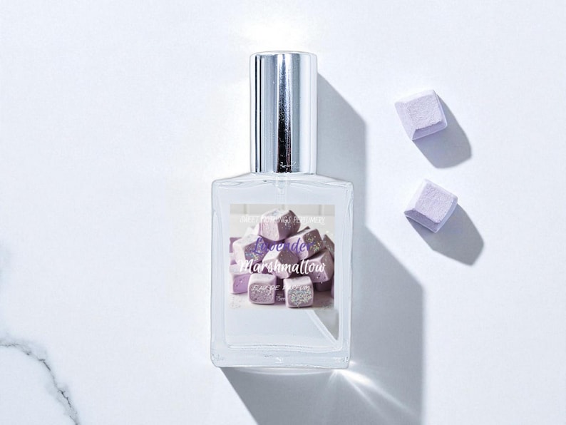 May include: A clear perfume bottle with a silver cap and a label featuring a graphic of purple marshmallow cubes. The text "Love de Marshmallow" is on the label. Two purple marshmallow cubes are next to the bottle.