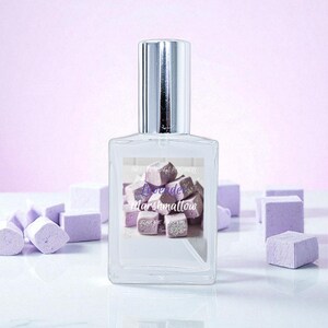 May include: A clear perfume bottle with a silver cap, featuring a label with the text "Love de Marshmallow" over a graphic of purple and white marshmallows. The background includes scattered marshmallow-shaped objects.