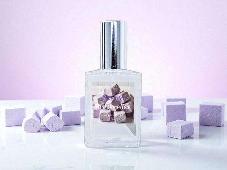May include: A clear perfume bottle with a silver cap, featuring a label with the text "Love de Marshmallow" over a graphic of purple and white marshmallows. The background includes scattered marshmallow-shaped objects.
