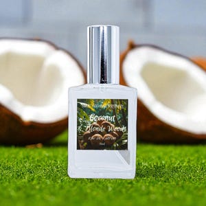 May include: A clear glass perfume bottle with a silver cap, labeled "Coconut & Blonde Woods Eau de Parfum 15ml." The bottle is centered on a green surface, with halved coconuts in the background. The label features a tropical design.