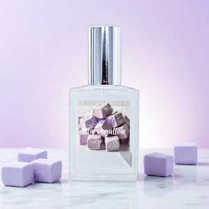 May include: A clear perfume bottle with a silver cap, featuring a label with the text "R&ecirc;ve de Marshmallow" and a graphic of purple marshmallow cubes. Several purple marshmallow cubes are scattered around the bottle on a marble surface.