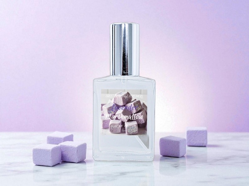 May include: A clear perfume bottle with a silver cap, featuring a label with the text "R&ecirc;ve de Marshmallow" and a graphic of purple marshmallow cubes. Several purple marshmallow cubes are scattered around the bottle on a marble surface.