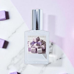 May include: A clear glass perfume bottle with a silver cap, featuring a label with the words "Love de Marshmallow" over a graphic of purple marshmallow cubes. Scattered around the bottle are more purple marshmallow cubes. The background is a mix of white marble and light purple.